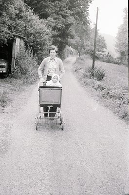 A woman in 1960s-era clothing pushes a vintage baby carriage along a rural gravel path, accompanied by a child seated inside....