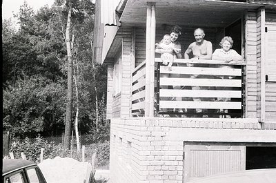 Three adults pose on a wooden balcony of a brick house, mid-20th century. Lush greenery and a parked car suggest a suburban o...