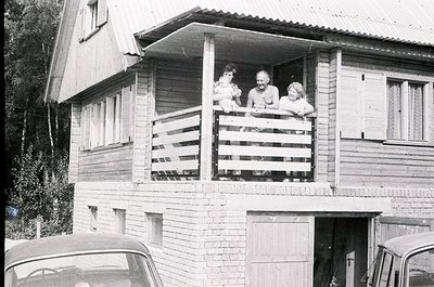 Three children pose on a wooden balcony of a mid-century brick-and-wood home, framed by horizontal railings. Classic 1960s fa...