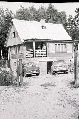 A 1960s-era wooden cabin with gabled roof and balcony, surrounded by dense forest. Two vintage cars (likely Lada or similar) ...