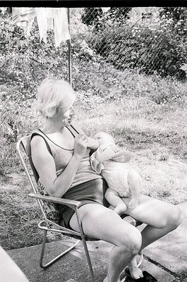 A woman in a sleeveless swimsuit cradles a small white dog in a metal folding chair, mid-1960s outdoor setting. Lush greenery...