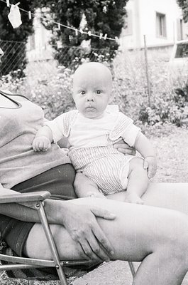 Black-and-white portrait of an infant seated in a wicker chair, mid-20th century. Baby wears a short-sleeve knit shirt and st...