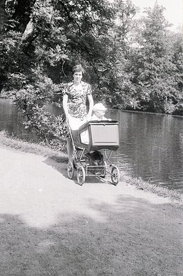 A woman in a patterned dress pushes a vintage baby carriage along a riverside path, mid-20th century. Lush greenery and water...