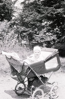 Vintage black-and-white photo of a child in a classic metal-framed pram on a forested path, mid-20th century. The pram’s bask...