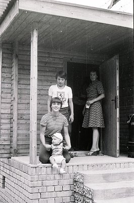 Family portrait on a brick porch, 1960s-1970s. Three individuals pose: a woman in floral dress holding a baby, a man in strip...