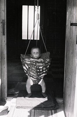 Vintage black-and-white photo of an infant suspended in a fabric swing, holding a pacifier, inside a rustic wooden doorway. S...