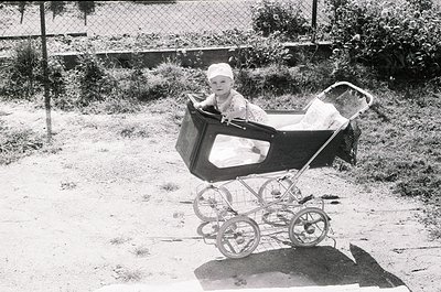 Vintage black-and-white photo of a child seated in a classic wicker pram with wire-spoked wheels, mid-20th century. Overgrown...