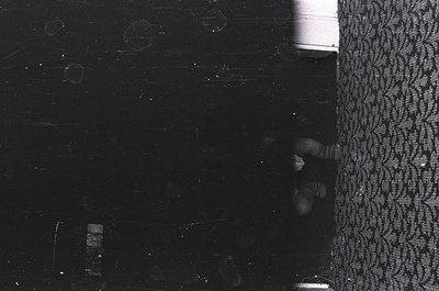 Vintage black-and-white photo of a child peeking from behind a patterned curtain, likely 1950s–1970s. Interior shows worn lin...