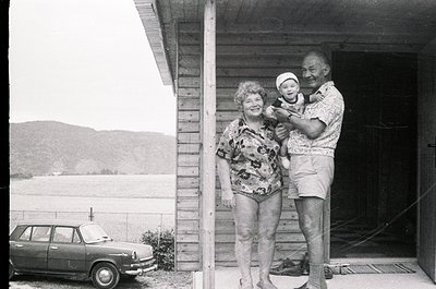 A mid-20th-century family portrait by a lakeside wooden cabin. Elderly couple (man in patterned shorts, woman in floral swims...