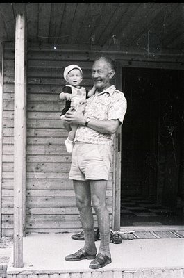 A mid-20th-century black-and-white photo of an elderly man holding a toddler on a wooden porch. The man wears a patterned shi...
