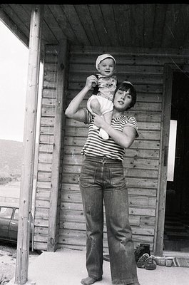 Black-and-white candid of a woman cradling a young child outside a rustic wooden structure, likely a rural home. She wears a ...