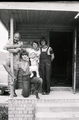 Family portrait in front of a rustic wooden structure, likely a barn or farmhouse porch, dated to the 1970s. Five individuals...