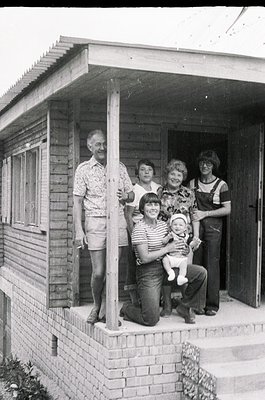 Family portrait in front of a wooden house, likely from the 1970s–1980s. Five adults and a child pose on a brick porch with a...