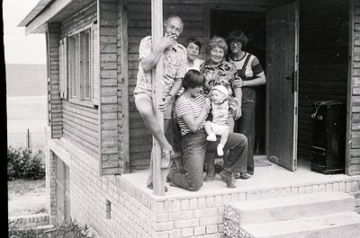 Family portrait on a brick porch of a 1970s-era wooden house, likely Eastern Bloc. Five individuals pose: adults in patterned...