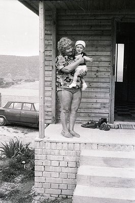 A woman in a floral swimsuit cradles a baby in a white cap on a wooden balcony, overlooking a coastal road. Classic car parke...