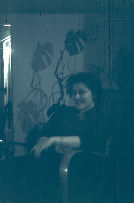 Vintage sepia-toned reflection of a woman in a dark coat and hat, seated indoors beside a wooden chair. Lighting suggests an ...