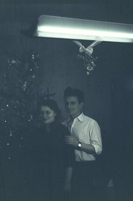 Black-and-white candid shot of two people standing under a fluorescent light, framed by a curtain of hanging ornaments. The m...