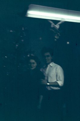 Vintage reflection shot of two people in a dimly lit indoor setting, likely a 1970s café or bar. The man wears a button-down ...