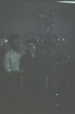 Vintage black-and-white photo of two people posing beside a decorated Christmas tree in an indoor setting, likely a home or s...