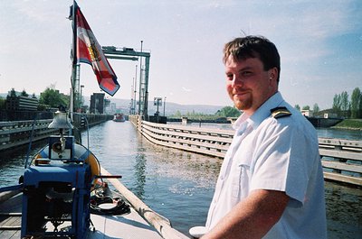 Maritime officer in white naval uniform poses near a canal lock, holding a rope. Industrial lock gate and crane in background...