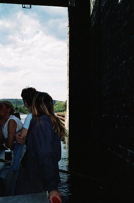 Two individuals stand indoors near a large wooden door, framed by natural light. One wears a light tank top and jeans, the ot...