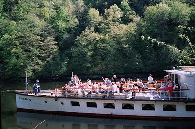 Vintage riverboat packed with mid-20th-century passengers enjoying a leisure cruise amid lush green forest. White hull with r...