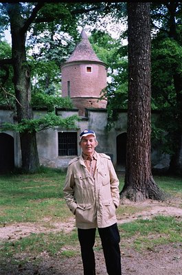 Man in vintage military-style jacket poses in front of a historic tower with conical roof and arched stone walls, surrounded ...
