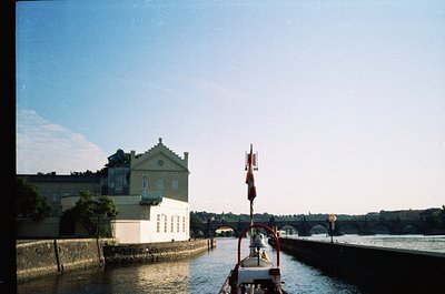 Vintage boat on calm river with classic European-style building in background, featuring pastel green walls and white trim. B...