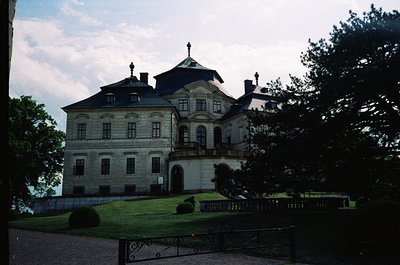 Neoclassical stone mansion with symmetrical façade, central balcony, and mansard roof. Ornate iron fence and manicured hedges...