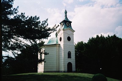 Neoclassical church tower with domed roof and clock, set against lush greenery. Light-colored stonework contrasts with dark t...