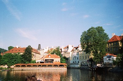 Historic European riverside scene with classic architecture—red-tiled roofs, pastel facades, and a stone bridge. Lush greener...
