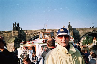 Vintage photo of Prague’s Charles Bridge with historic Gothic towers and statues. Elderly man in sunglasses and cap poses nea...