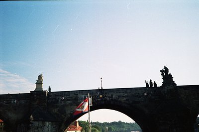 Historic stone bridge with Gothic statues and arch framing a flagpole displaying Czech and EU flags. Urban landscape with dis...