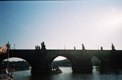 Historic stone bridge with Gothic statues over the Vltava River, likely Prague’s Charles Bridge. Sunlight casts long shadows ...