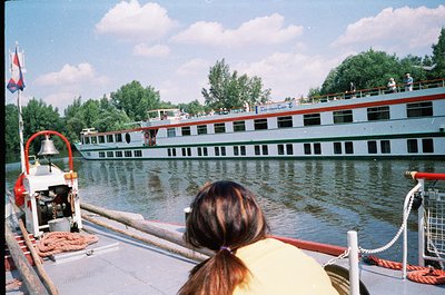 Vintage river cruise scene featuring a mid-20th century passenger boat docked alongside a larger vessel. The foreground shows...