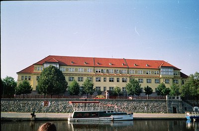 Three-story yellow building with red-tiled gable roofs, symmetrical windows, and decorative brickwork near waterfront. Stone ...