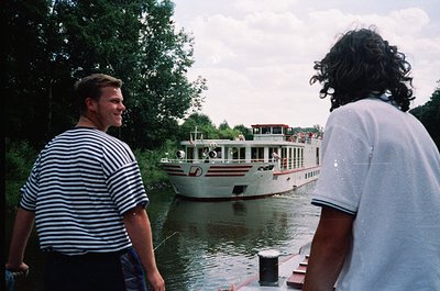 Two men stand on a dock beside a river, observing a white passenger boat with red accents and a flag. The man on the left wea...
