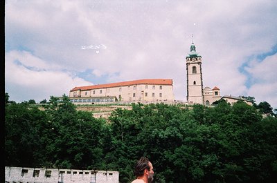 Historic fortress complex perched atop a hill, featuring a prominent bell tower and red-tiled rooftops. Lush greenery surroun...