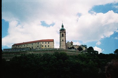 Historic fortress complex atop a hill, featuring a prominent bell tower with a green spire and adjacent yellowish stone build...
