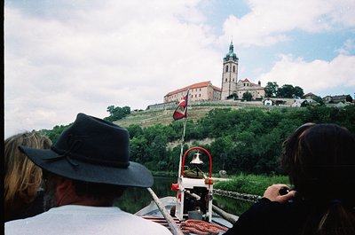 Boat tour view of medieval fortress with bell tower atop hillside, featuring red-and-white flag. Foreground shows passengers ...