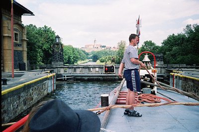 Lockkeeper operating a canal lift bridge, wearing striped shirt and holding a rope bell. Historic European town with castle r...