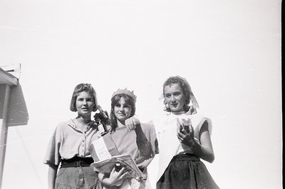 Three young women in 1950s-style dresses pose outdoors, likely at a school event or festival. Center figure wears a crown and...