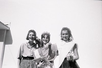 Three women pose outdoors in mid-20th century attire, likely 1950s–1960s. Center figure wears a paper crown and sash, suggest...
