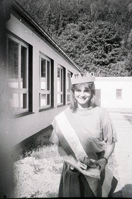 Young woman in graduation regalia poses outside a utilitarian building, likely a school or dormitory, with forested hills in ...