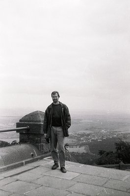 Black-and-white portrait of a man in 1970s-era attire (leather jacket, rolled-up trousers) standing atop a stone terrace with...
