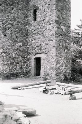 Medieval stone tower with small arched doorway and narrow slit window, surrounded by scattered rubble and logs. Likely a hist...