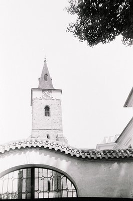 Mid-century church tower with pointed steeple and arched bell window, framed by ornate wrought-iron gate. Stone façade and ti...