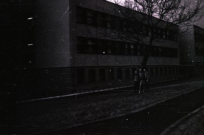 Mid-century brutalist concrete building with vertical window bands, captured in monochrome. Two figures in dark coats walk al...