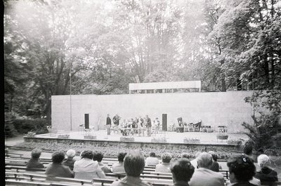 Mid-century outdoor amphitheater with tiered wooden seating under dense trees. Stage features minimalist concrete platform wi...