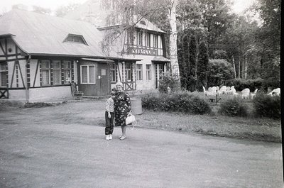 Mid-20th century black-and-white photo of a woman in traditional attire holding a basket, standing near a sign reading "Kavar...
